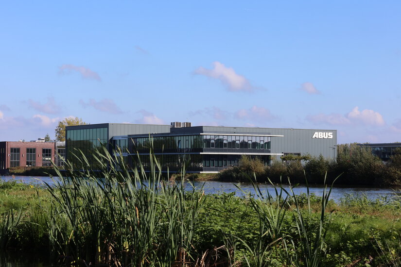 Zicht op het moderne bedrijfsgebouw van ABUS Kraansystemen B.V. in Nederland, omgeven door groene vegetatie en een meer, met strakke lijnen en grote glazen gevels